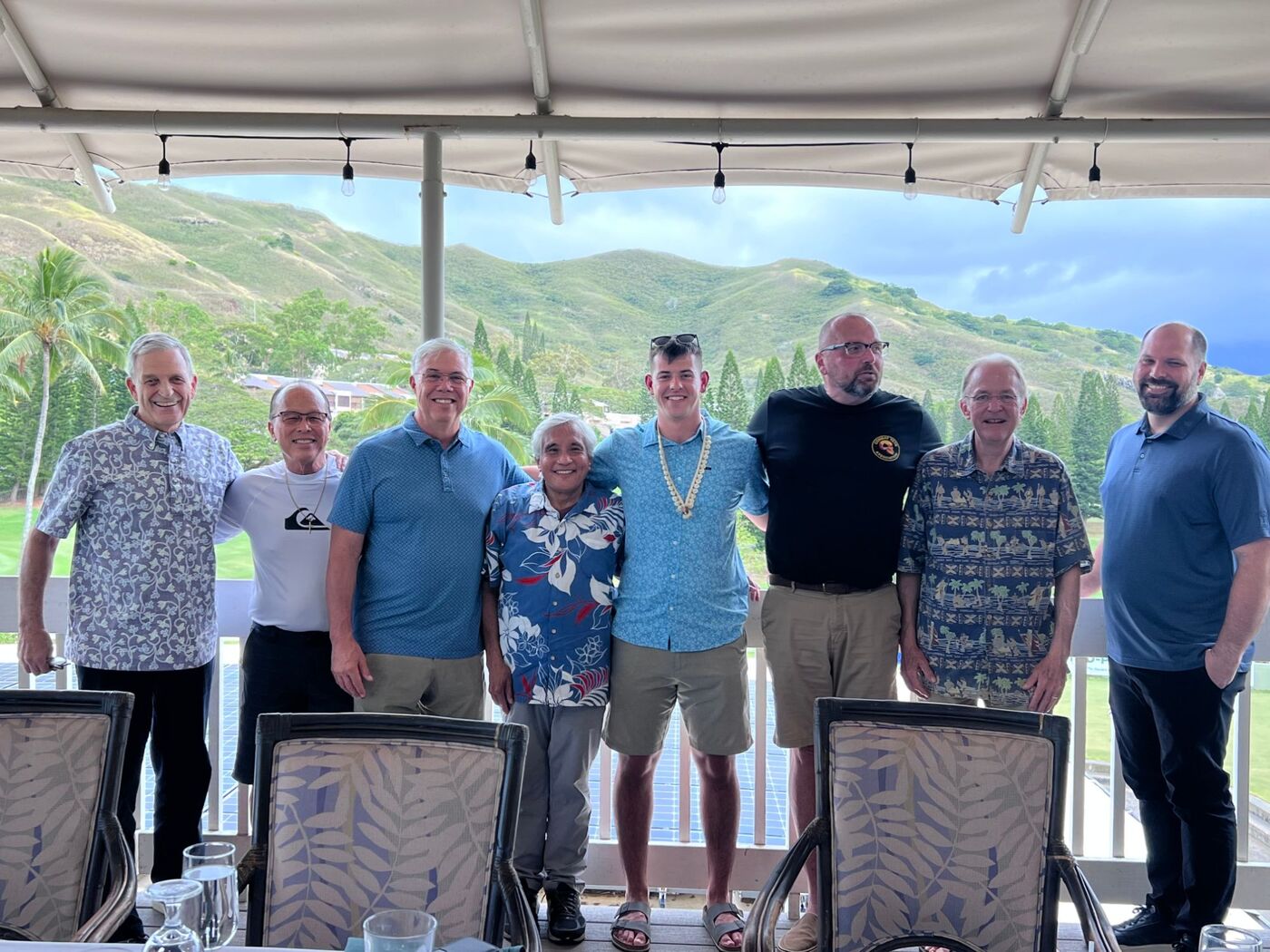 Eight men of the fellowship on a lanai, with green Koolau ridges behind them